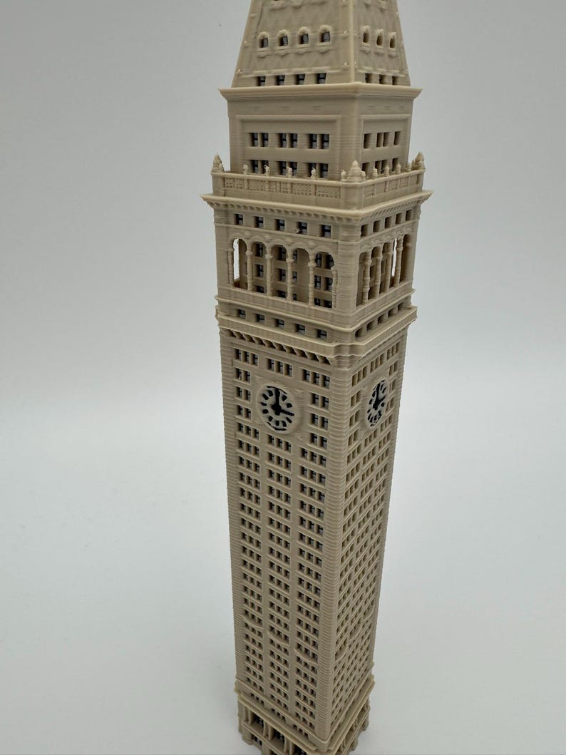 May include: A beige, three-dimensional printed model of a tall clock tower building. The structure features numerous rectangular windows, a prominent clock face on two visible sides, and an ornate, multi-tiered top section with arched openings.