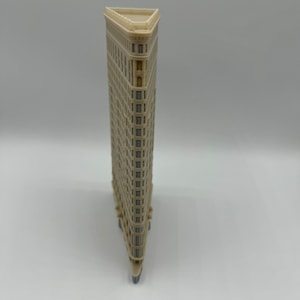 May include: A beige miniature model of the Flatiron Building in New York City. The model is made of plastic and has a triangular shape. The building has many windows and a flat roof.