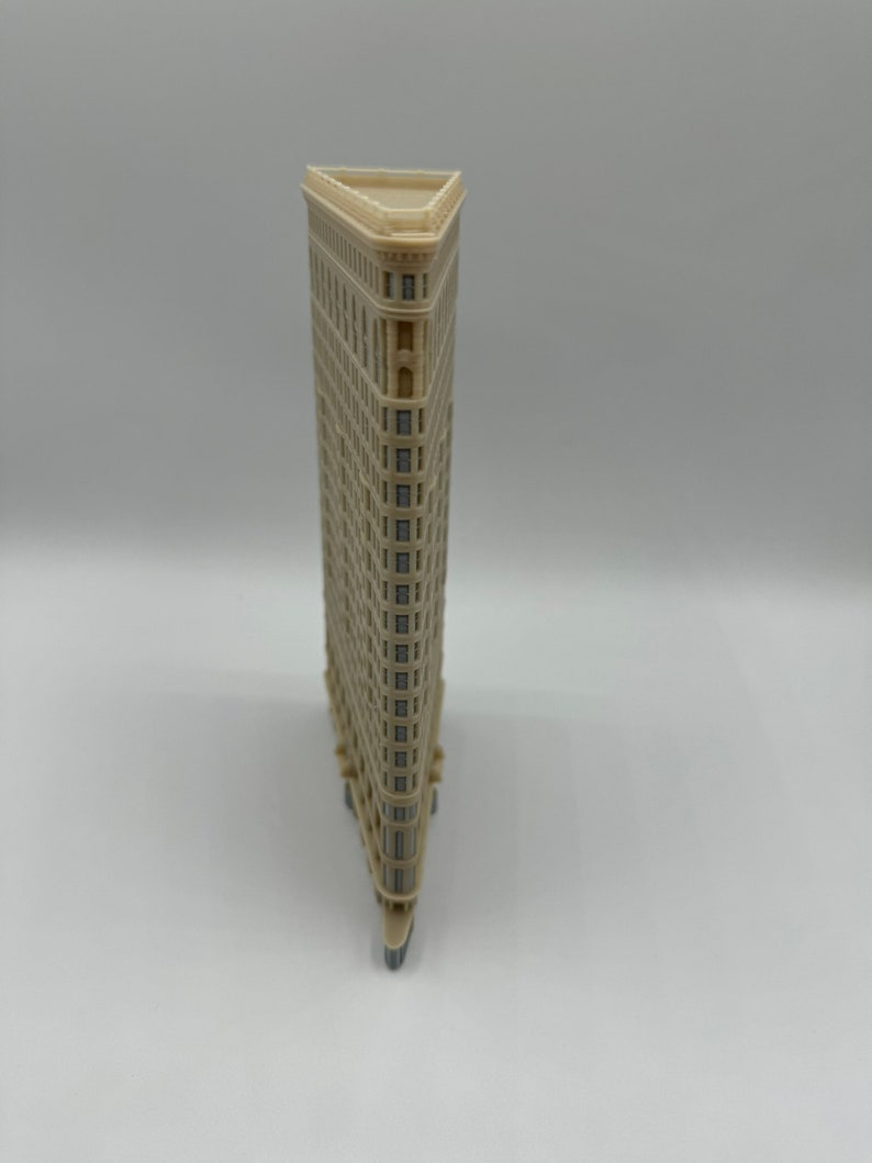 May include: A beige miniature model of the Flatiron Building in New York City. The model is made of plastic and has a triangular shape. The building has many windows and a flat roof.
