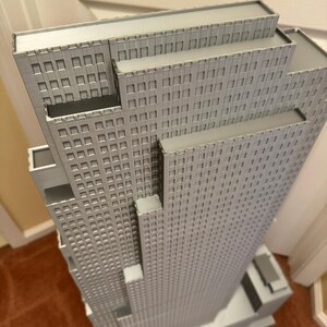 May include: A detailed, gray architectural model of a skyscraper. The model features multiple tiers and a grid-like pattern of windows across its facade. The building is set on a rectangular base.