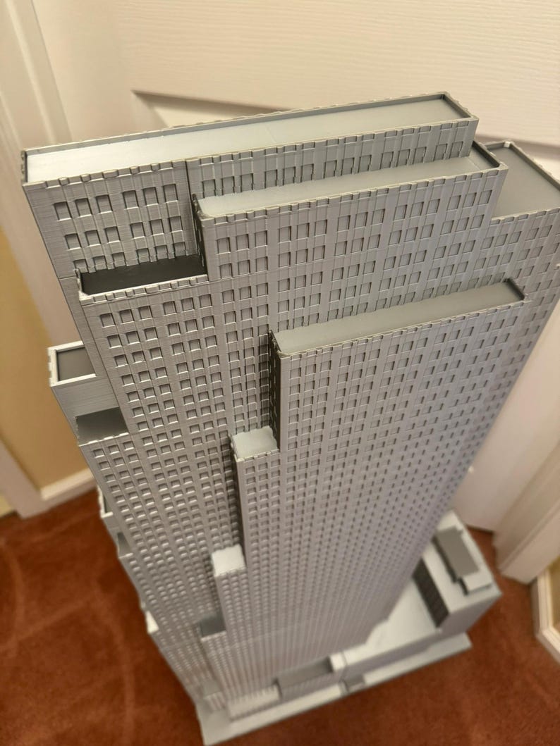 May include: A detailed, gray architectural model of a skyscraper. The model features multiple tiers and a grid-like pattern of windows across its facade. The building is set on a rectangular base.