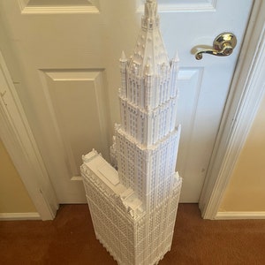 May include: A white 3D printed model of a tall skyscraper with intricate details and a pointed spire.