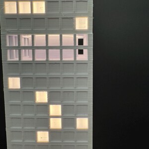 May include: A white, 3D-printed model of a high-rise building with illuminated windows. The windows glow with a warm, yellow light, contrasting against the white structure. The building's design features a grid-like pattern of windows.