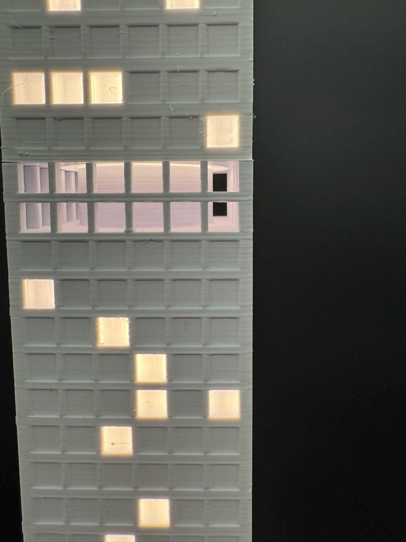 May include: A white, 3D-printed model of a high-rise building with illuminated windows. The windows glow with a warm, yellow light, contrasting against the white structure. The building's design features a grid-like pattern of windows.