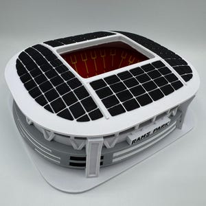 Pode incluir: Modelo impresso em 3D de um estádio, com exterior branco e painéis pretos no telhado. O estádio tem uma base cinza e o texto "RAMS PARK" é visível. O interior é vermelho com detalhes amarelos.