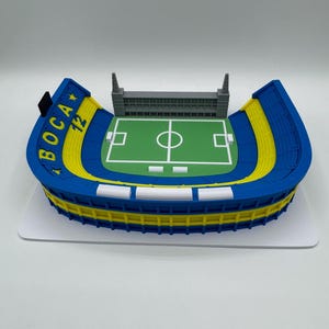 May include: A blue, yellow, and white stadium model with the text "BOCA 12". The model has a green pitch with white markings and a grey structure at the top. The stadium sits on a white base.
