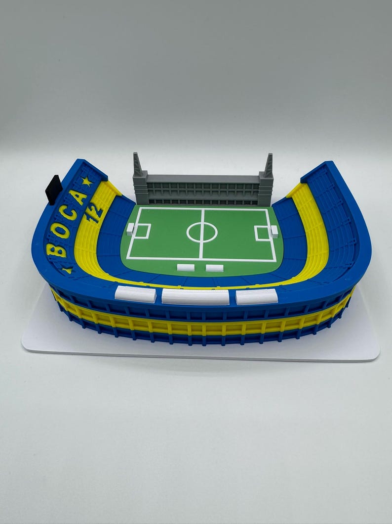 May include: A blue, yellow, and white stadium model with the text "BOCA 12". The model has a green pitch with white markings and a grey structure at the top. The stadium sits on a white base.