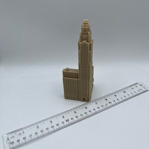 May include: A beige 3D printed model of a tall skyscraper with a pointed roof. The model is about 6 inches tall and is sitting on a white surface next to a ruler.