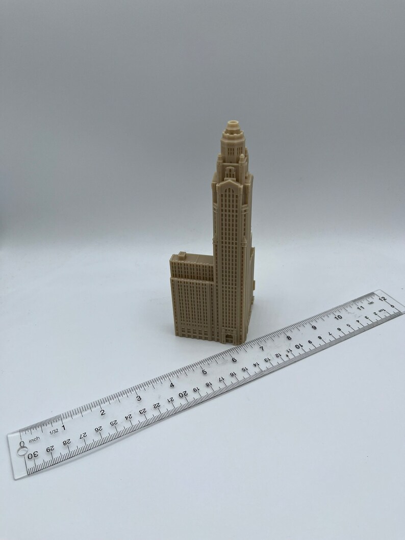 May include: A beige 3D printed model of a tall skyscraper with a pointed roof. The model is about 6 inches tall and is sitting on a white surface next to a ruler.