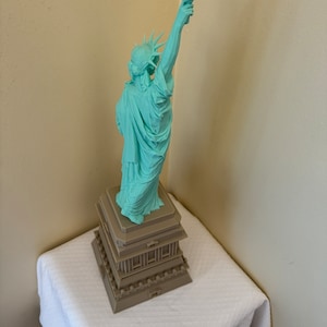 May include: A teal green statue of the Statue of Liberty with a brown base. The statue is standing on a white tablecloth.
