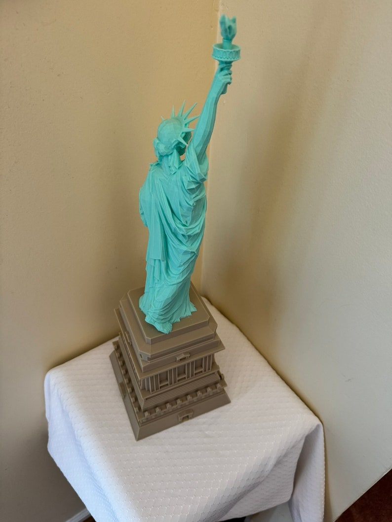 May include: A teal green statue of the Statue of Liberty with a brown base. The statue is standing on a white tablecloth.