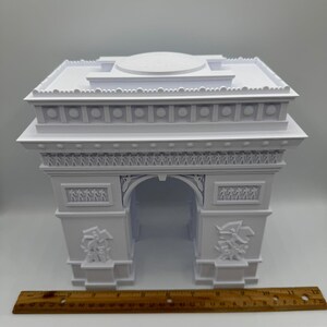 Może przedstawiać: Biały model architektoniczny Łuku Triumfalnego z precyzyjnymi rzeźbami. Model ma około 25 cm szerokości, z linijką umieszczoną poniżej dla skali. Łuk posiada centralny otw&oacute;r i dekoracyjne rzeźby reliefowe.