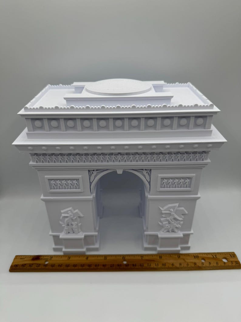 Może przedstawiać: Biały model architektoniczny Łuku Triumfalnego z precyzyjnymi rzeźbami. Model ma około 25 cm szerokości, z linijką umieszczoną poniżej dla skali. Łuk posiada centralny otw&oacute;r i dekoracyjne rzeźby reliefowe.