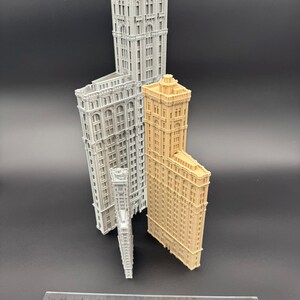 May include: A selection of miniature building models in different sizes and colours. The models include a light grey, a tan, and a white building. A ruler is placed at the bottom of the image.