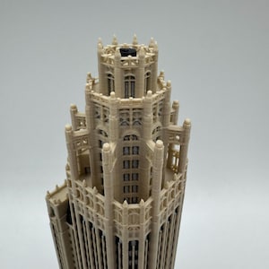 May include: A beige 3D printed architectural model of a skyscraper featuring intricate gothic-style detailing, vertical columns, and a tiered crown structure with a small black circular component visible at the very top center of the building.