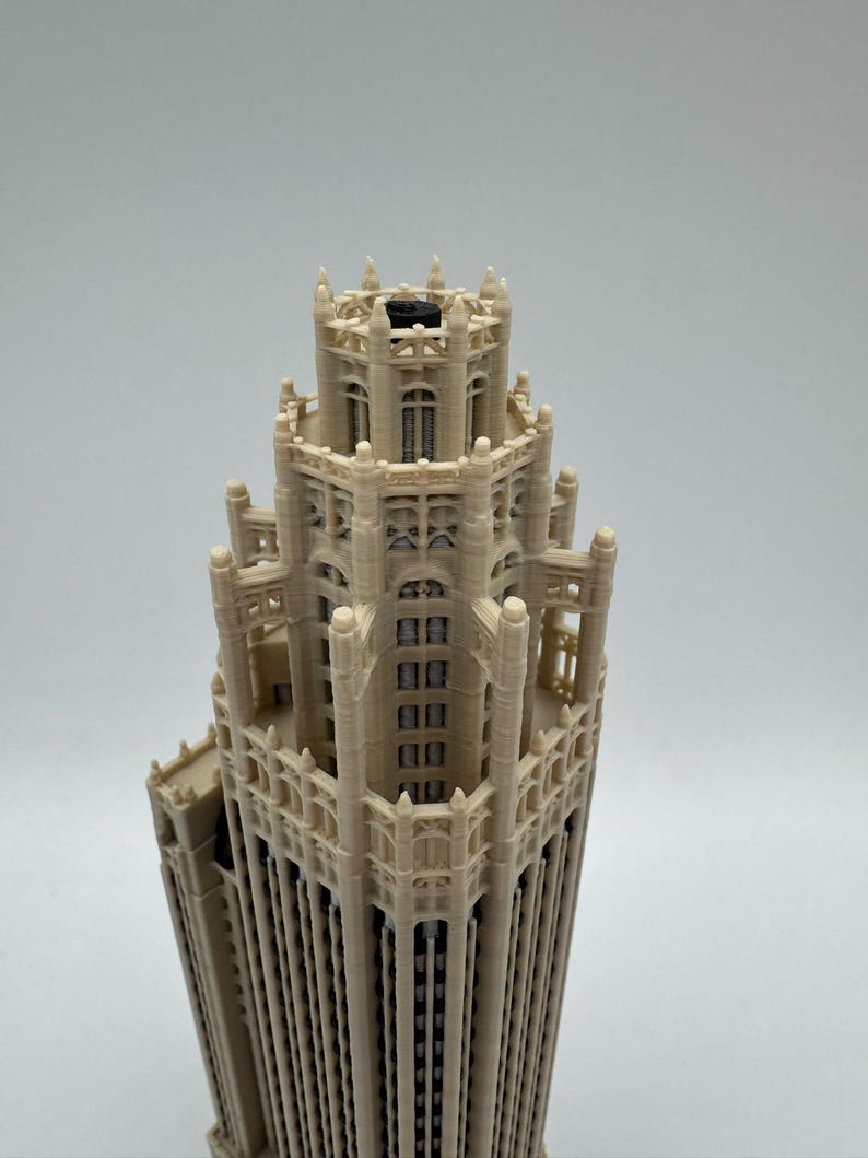 May include: A beige 3D printed architectural model of a skyscraper featuring intricate gothic-style detailing, vertical columns, and a tiered crown structure with a small black circular component visible at the very top center of the building.