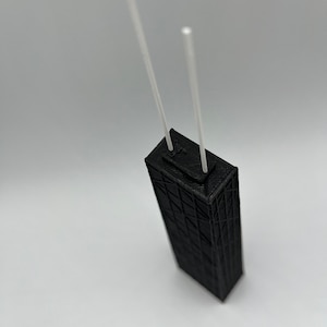 May include: A black, 3D-printed model of a skyscraper with two white, vertical rods extending from the top. The building has a detailed, geometric facade. The model is set against a plain white background.