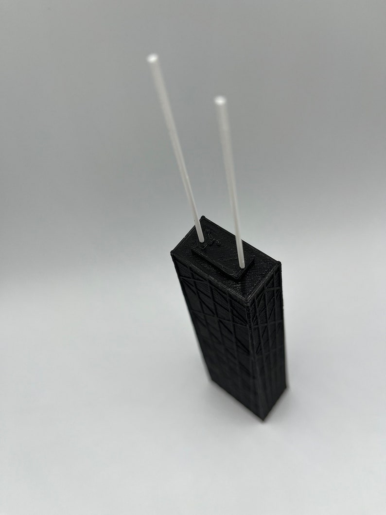 May include: A black, 3D-printed model of a skyscraper with two white, vertical rods extending from the top. The building has a detailed, geometric facade. The model is set against a plain white background.
