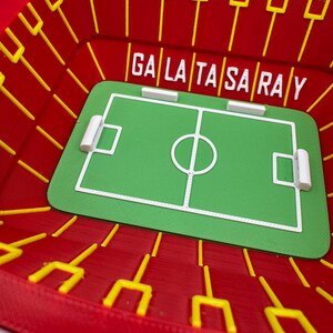 May include: A miniature stadium model in red with yellow goalposts and the text "GALATASARAY". The stadium features a green soccer field with white markings and goal areas. A unique decorative item.