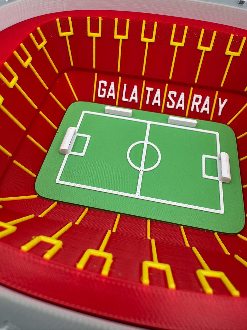 May include: A miniature stadium model in red with yellow goalposts and the text "GALATASARAY". The stadium features a green soccer field with white markings and goal areas. A unique decorative item.