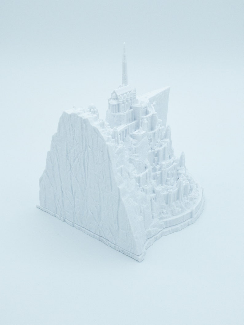 May include: A white 3D printed model of a castle on a cliff. The castle has a tall spire and many towers. The cliff is textured to look like rock.