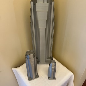 May include: Three 3D printed models of skyscrapers in a silver gray color. The tallest building is about 12 inches tall, the middle building is about 6 inches tall, and the smallest building is about 4 inches tall.