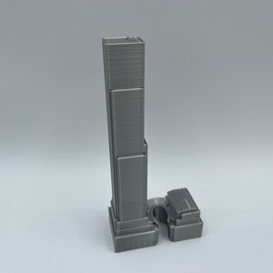 May include: A gray 3D printed model of a skyscraper with a rectangular base and a tall, narrow tower.
