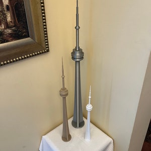 May include: Three miniature CN Tower models in varying sizes and colors. The tallest is silver, the middle one is tan, and the smallest is white. They are displayed on a white cloth-covered surface, set against a neutral-toned wall.
