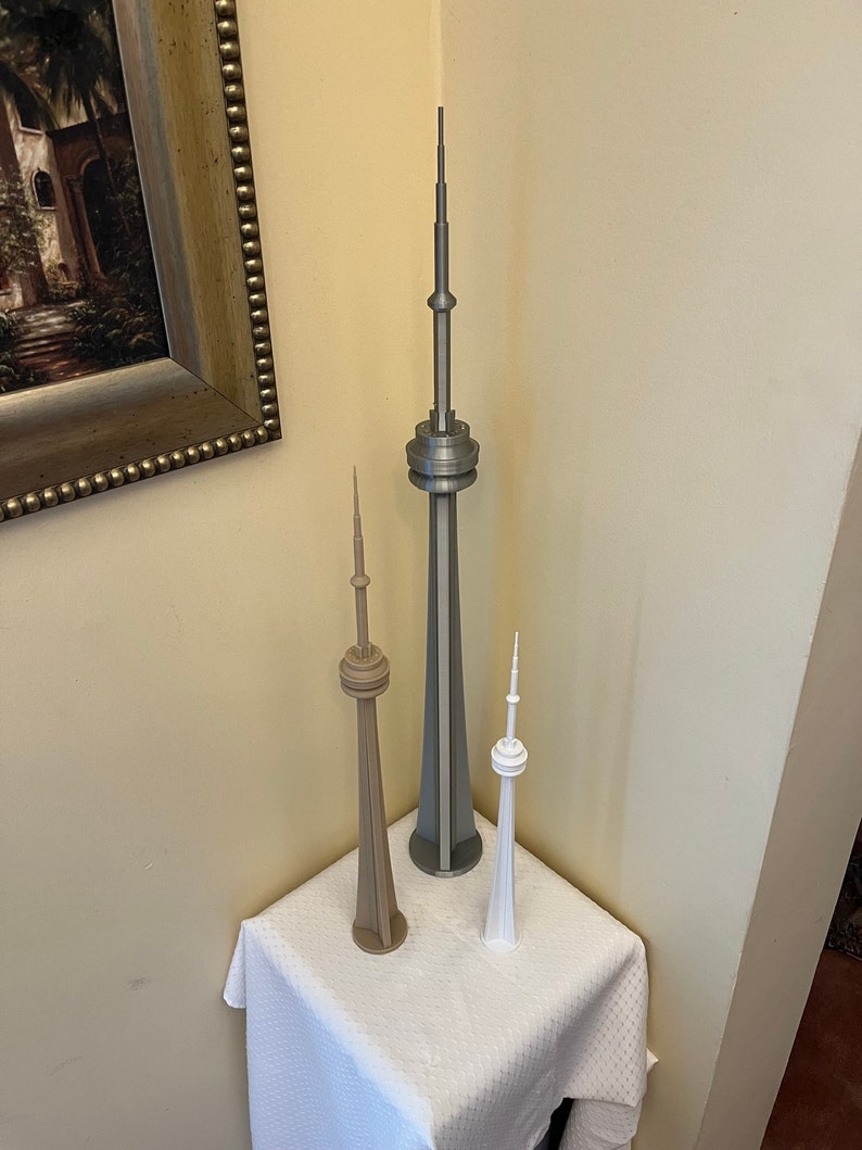 May include: Three miniature CN Tower models in varying sizes and colors. The tallest is silver, the middle one is tan, and the smallest is white. They are displayed on a white cloth-covered surface, set against a neutral-toned wall.