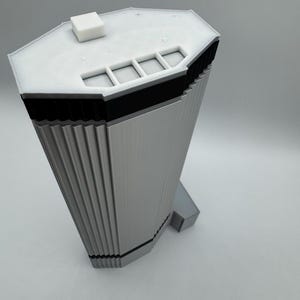 May include: A 3D-printed model of a modern skyscraper. The building is primarily light gray with black accents and a white roof. The model has a unique, angular design with vertical lines and a small square structure on top.
