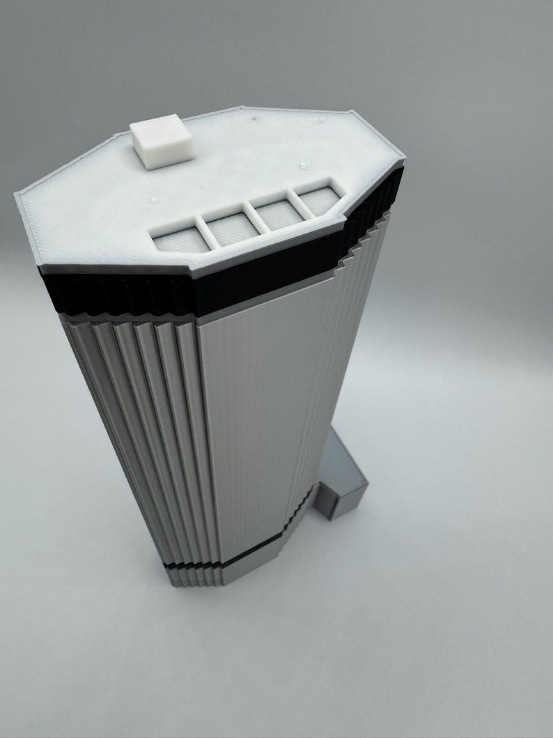 May include: A 3D-printed model of a modern skyscraper. The building is primarily light gray with black accents and a white roof. The model has a unique, angular design with vertical lines and a small square structure on top.