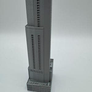 May include: A gray, 3D-printed model of a skyscraper. The building has a rectangular base with detailed architectural features. The tower rises with multiple rows of windows, showcasing a modern design. The model is set against a plain background.