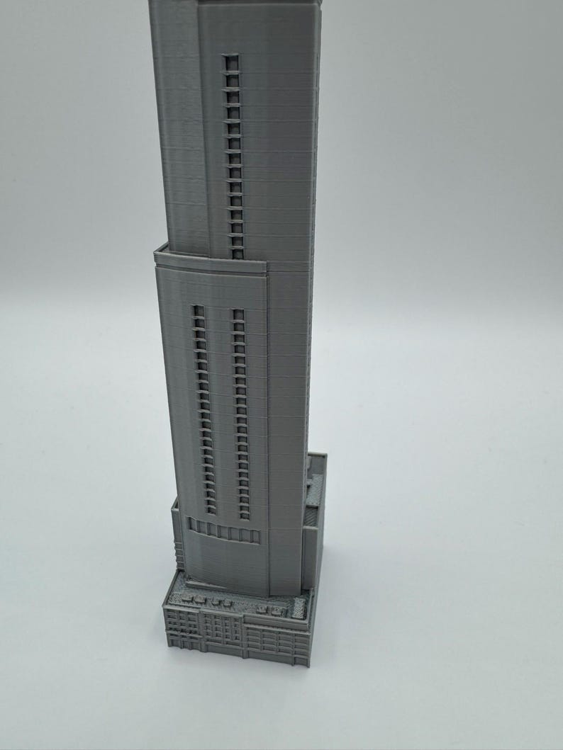 May include: A gray, 3D-printed model of a skyscraper. The building has a rectangular base with detailed architectural features. The tower rises with multiple rows of windows, showcasing a modern design. The model is set against a plain background.