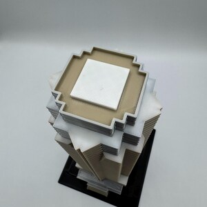 May include: A 3D-printed geometric sculpture with a pixelated design. The sculpture features stacked square layers in beige, white, and gray, with a black base. The top layer has a white square in the center.