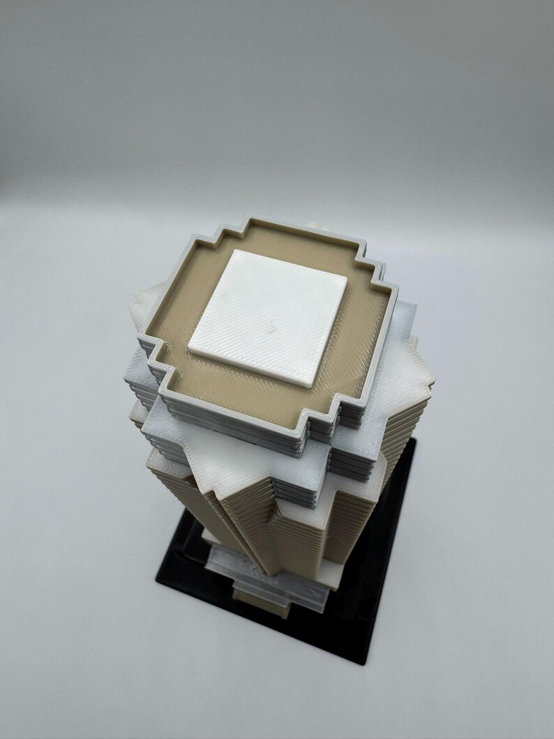 May include: A 3D-printed geometric sculpture with a pixelated design. The sculpture features stacked square layers in beige, white, and gray, with a black base. The top layer has a white square in the center.