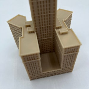 May include: A beige 3D printed model of a tall skyscraper building with a square base and a rectangular opening in the center.