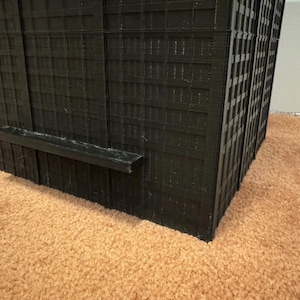 May include: A black, 3D-printed model of a skyscraper. The building features a grid of windows and a flat, rectangular base. The model is placed on a brown carpet.