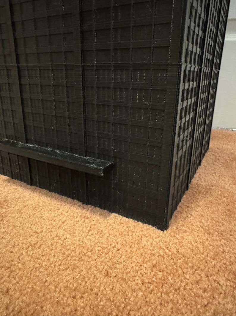 May include: A black, 3D-printed model of a skyscraper. The building features a grid of windows and a flat, rectangular base. The model is placed on a brown carpet.