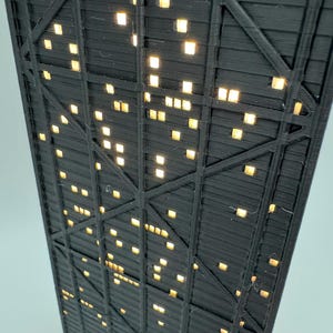 May include: Low-angle view of a black, geometric structure with numerous square, illuminated windows. The structure has a grid-like pattern with diagonal and horizontal supports. The windows emit a warm, yellow light, creating a contrast against the dark framework.