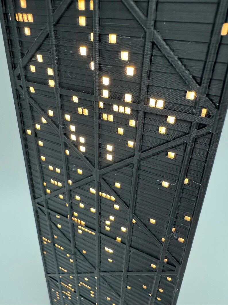 May include: Low-angle view of a black, geometric structure with numerous square, illuminated windows. The structure has a grid-like pattern with diagonal and horizontal supports. The windows emit a warm, yellow light, creating a contrast against the dark framework.