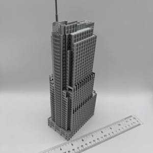 May include: A gray miniature skyscraper model with a tall antenna. The building has many windows and architectural details. A clear ruler with inch markings is placed next to the building.