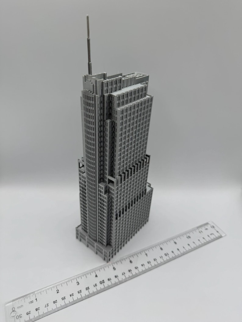 May include: A gray miniature skyscraper model with a tall antenna. The building has many windows and architectural details. A clear ruler with inch markings is placed next to the building.
