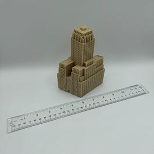 May include: A tan-colored miniature building model with a detailed architectural design. A clear ruler with inch measurements is placed next to the model. The model appears to be approximately 4 inches tall.