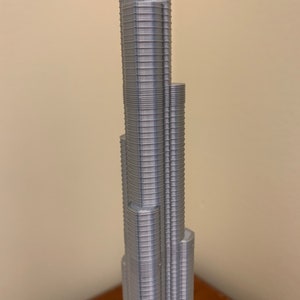 May include: A silver 3D printed model of a skyscraper. The model is detailed with many small, horizontal lines that represent the building's floors.