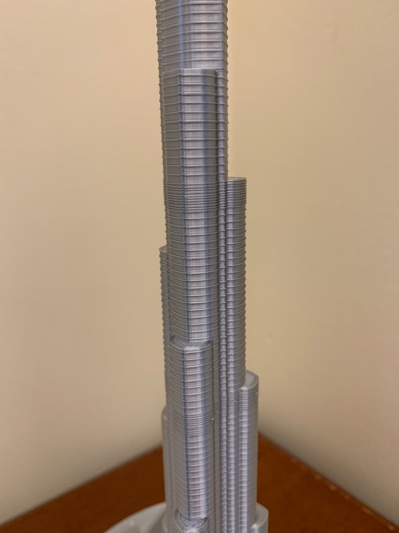 May include: A silver 3D printed model of a skyscraper. The model is detailed with many small, horizontal lines that represent the building's floors.
