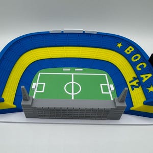 May include: A miniature stadium model featuring blue and yellow seating, a green pitch, and grey structures. The text "BOCA 12" is visible on the blue section. The model is on a white base.
