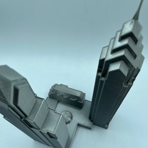 May include: A 3D printed model of two skyscrapers, one taller than the other, with a small plaza between them. The model is made of gray plastic and has a detailed design.