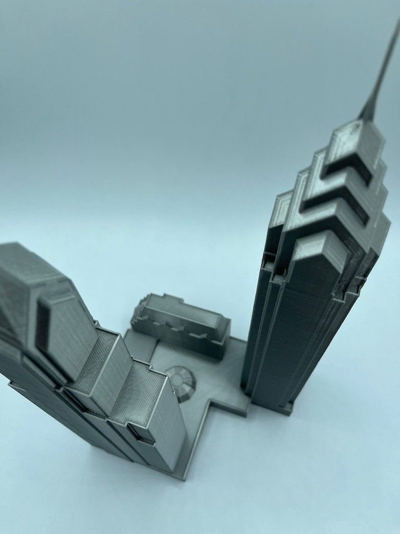 May include: A 3D printed model of two skyscrapers, one taller than the other, with a small plaza between them. The model is made of gray plastic and has a detailed design.