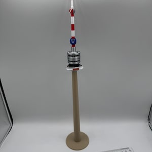 Op de afbeelding: Een miniatuurmodel van een toren met een beige basis en paal, een zilver-grijs middengedeelte en een rood, wit en blauw topje. Een liniaal is onderaan de afbeelding geplaatst.