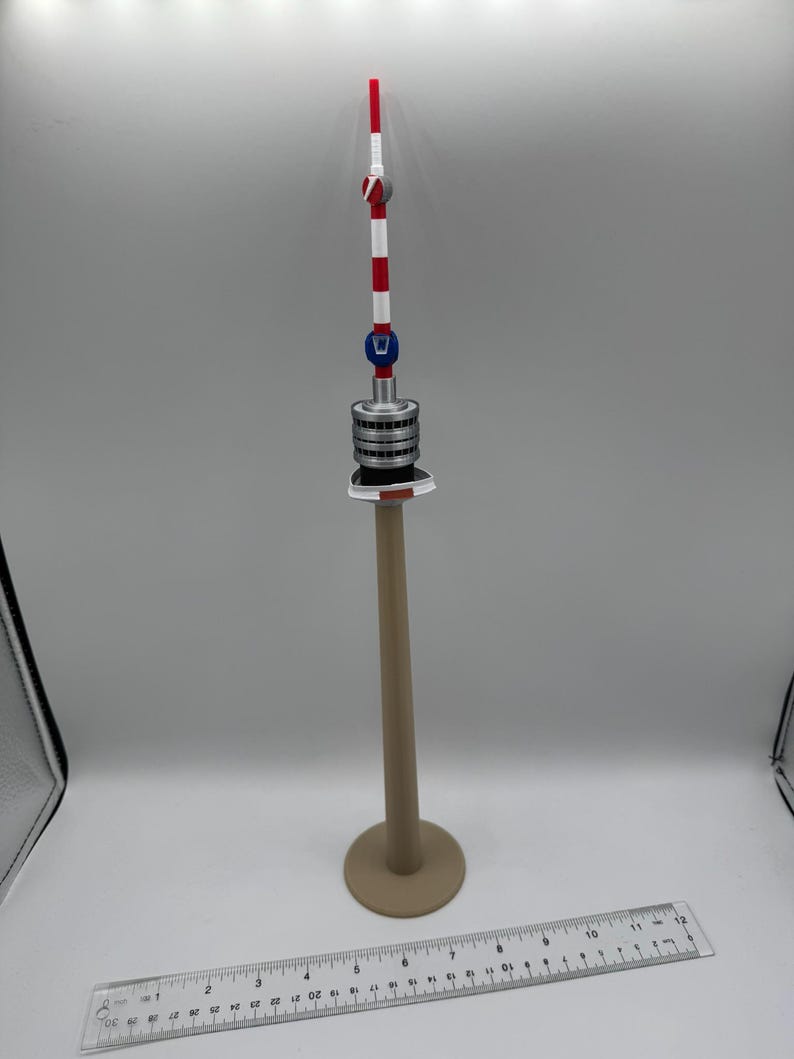 Op de afbeelding: Een miniatuurmodel van een toren met een beige basis en paal, een zilver-grijs middengedeelte en een rood, wit en blauw topje. Een liniaal is onderaan de afbeelding geplaatst.